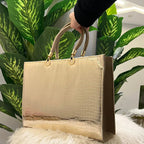 Beige handbag with gold handles held by a hand against a plant and white surface background