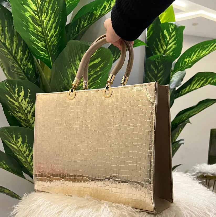 Beige handbag with gold handles held by a hand against a plant and white surface background