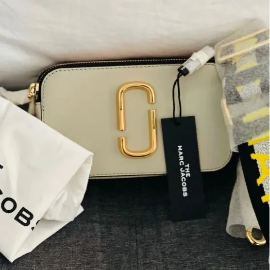 Beige handbag with gold clasp and visible Marc Jacobs tag on a dark surface.