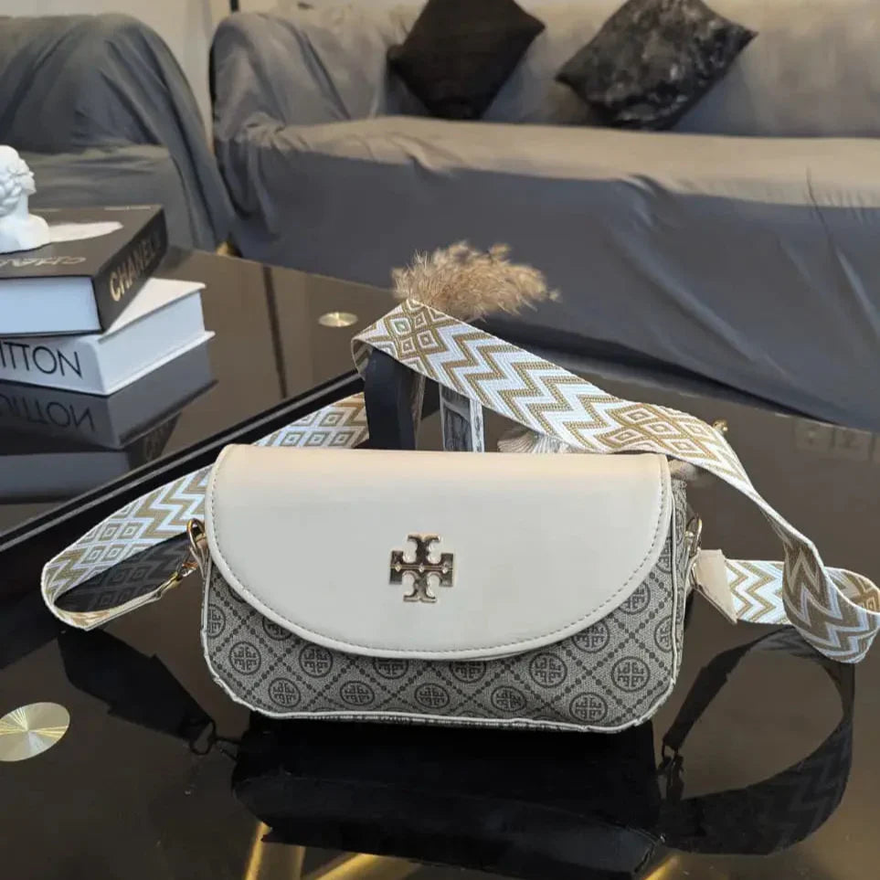 White handbag with patterned strap on a reflective surface, with books and a couch in the background.