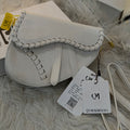 White handbag with braided handle on a fluffy white surface, featuring a QINGMUXI tag.